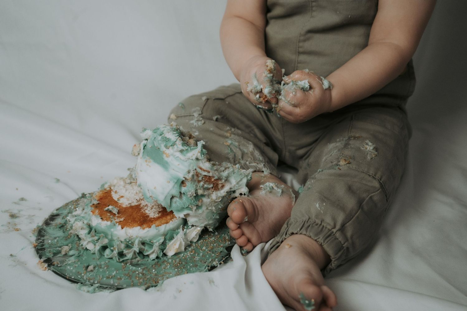 Baby in olive green overalls playing with blue frosting or paint on white surface.