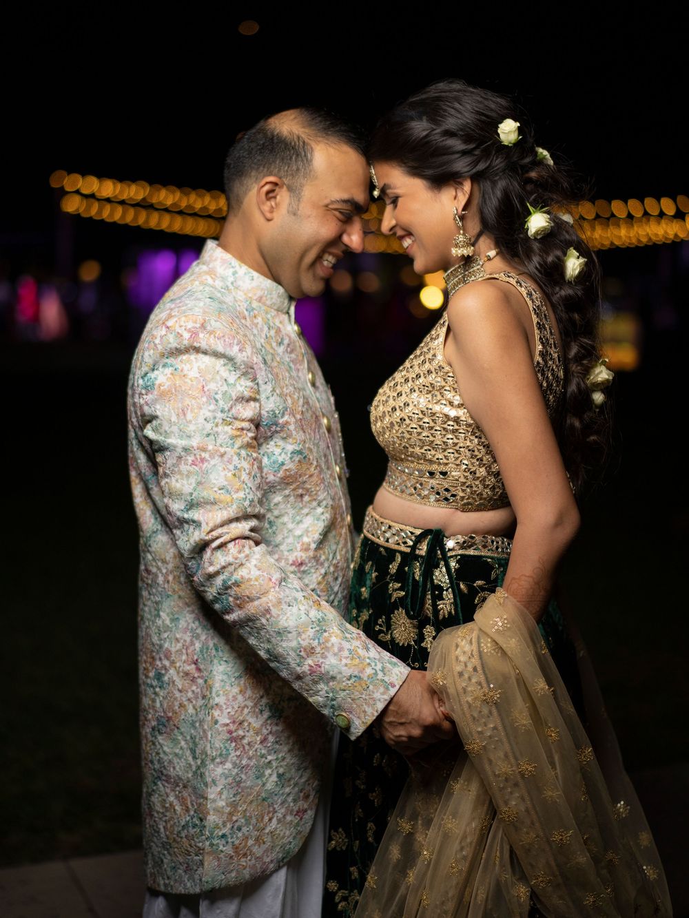 Indian couple in traditional wedding attire sharing a romantic moment at night, man in floral sherwani, woman in gold lehenga.