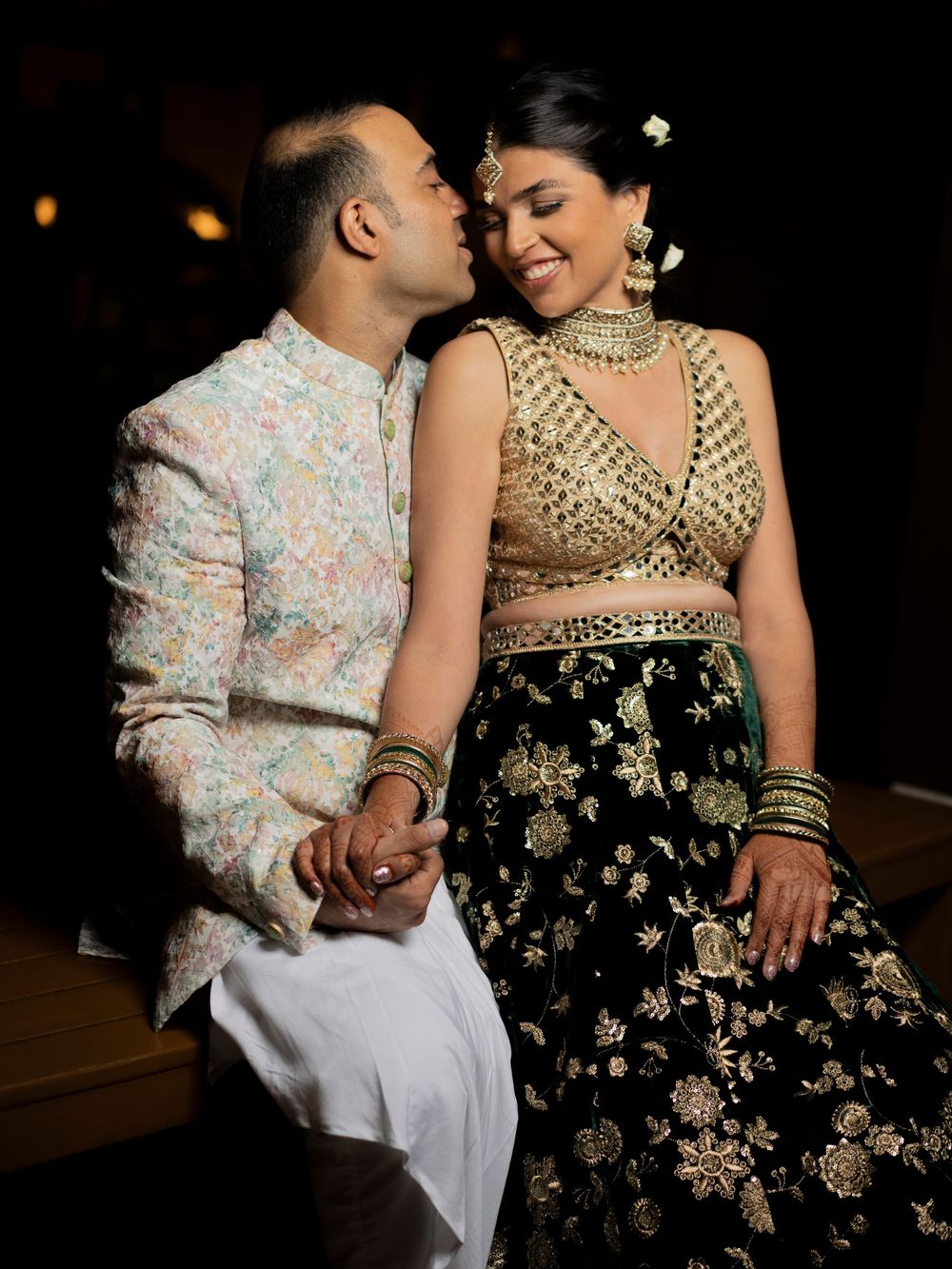 Indian couple in traditional attire sharing a romantic moment, bride in gold and black lehenga, groom in floral sherwani.