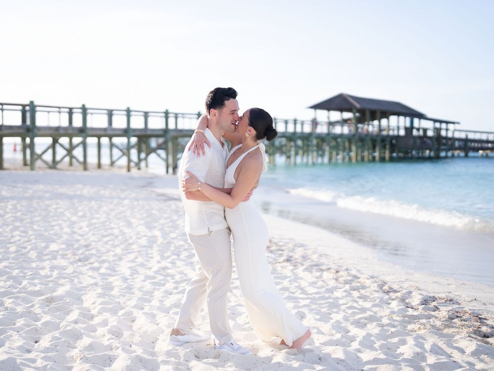 Couple sharing a romantic kiss on a white sand beach near a wooden pier on their wedding day.