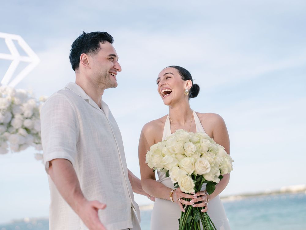 Couple laughing together on beach, bride holding white rose bouquet in elegant halter dress, groom in white linen.