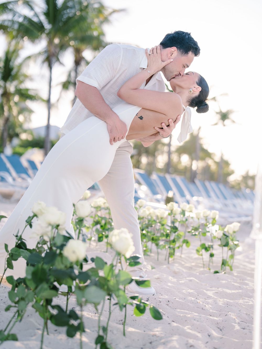 Romantic beach proposal dip kiss among white roses in sand, couple dressed in white with palm trees backdrop.