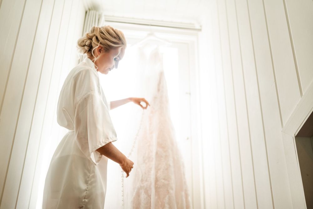 Bride in white robe admiring her lace wedding dress hanging in a bright, sunlit room on her wedding day.