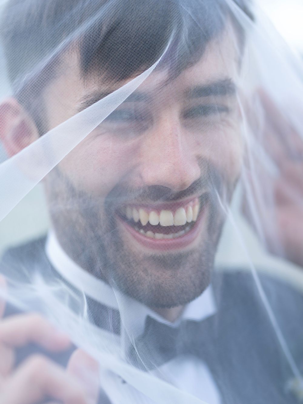 Smiling groom in a tuxedo laughing joyfully beneath a flowing white bridal veil on his wedding day.