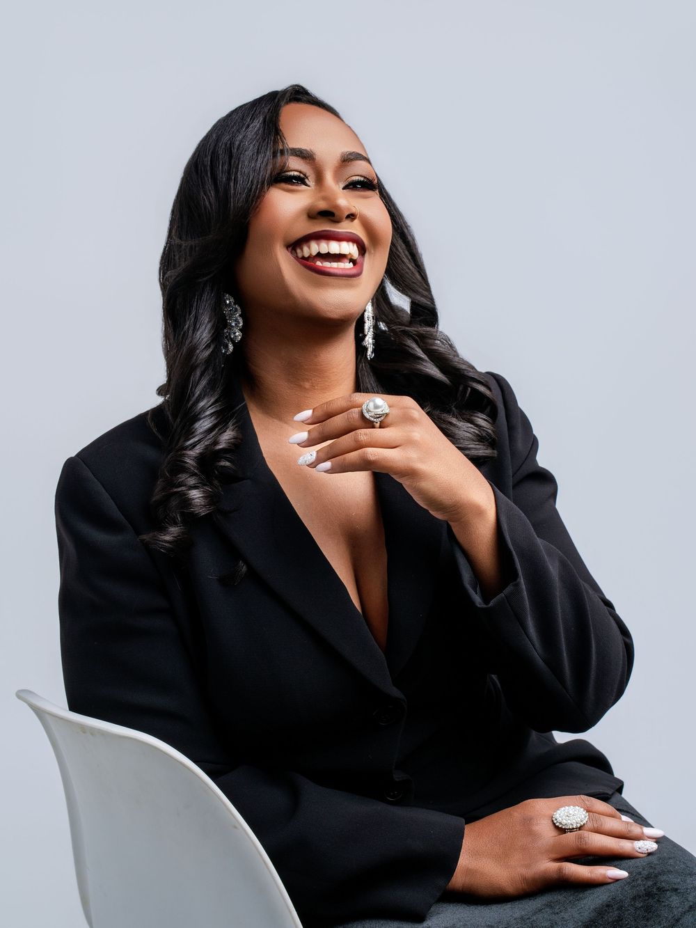 Smiling woman in black blazer sitting gracefully, wearing elegant drop earrings and rings, with long wavy hair.
