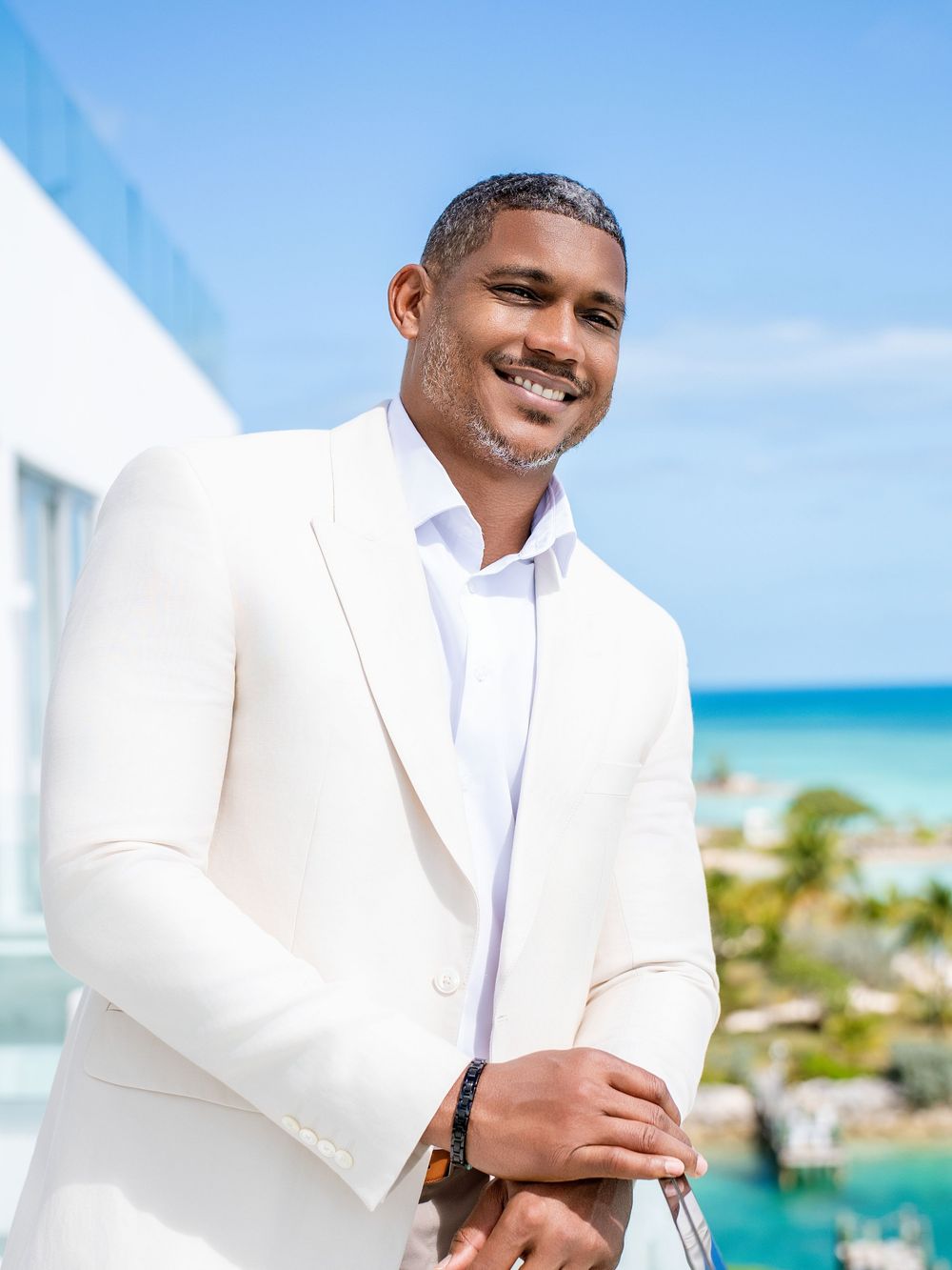 Professional wearing a white suit jacket poses against a tropical beach backdrop with turquoise waters and blue sky.