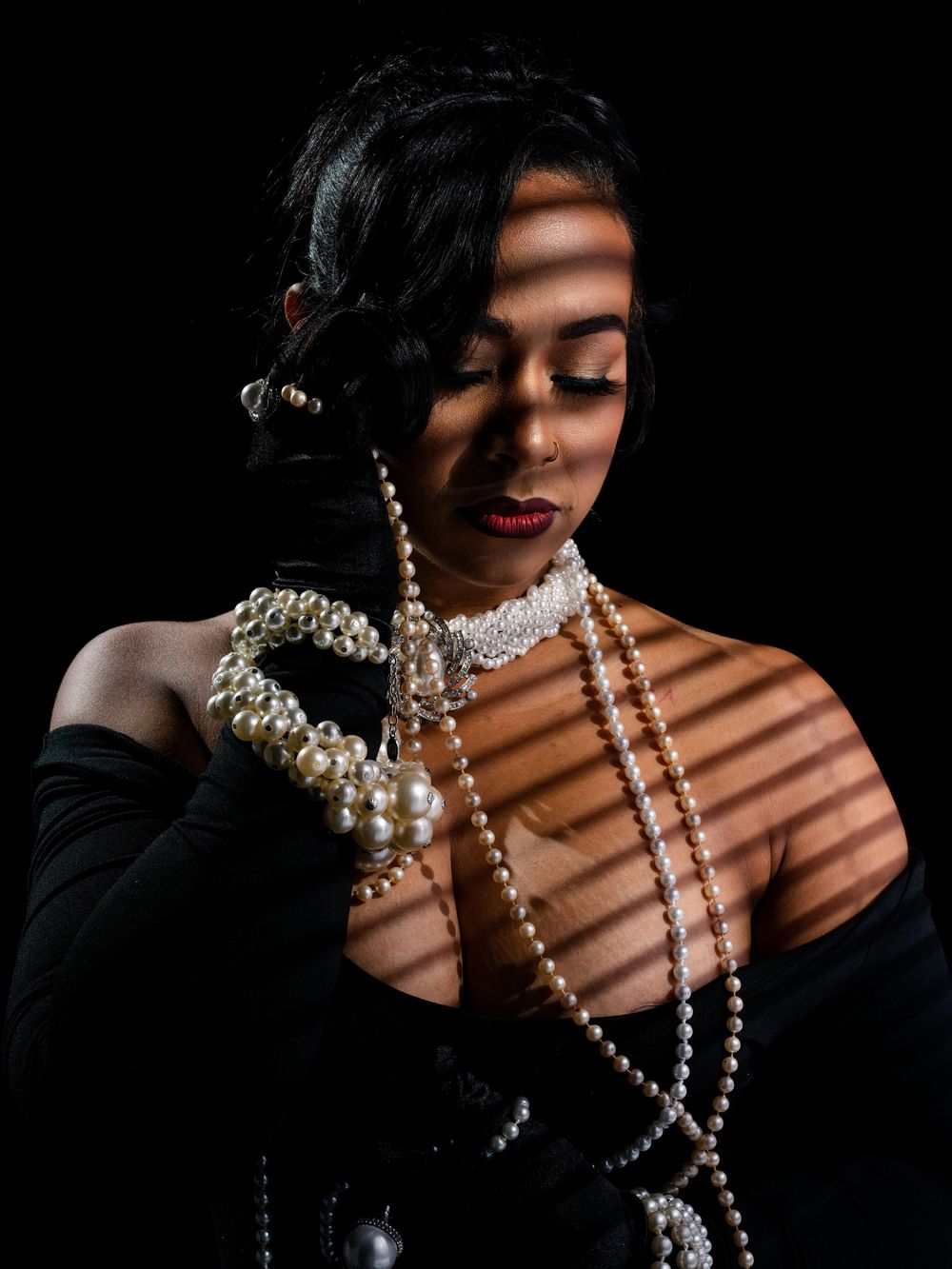 Elegant woman in black gloves holding pearl necklaces with dramatic light stripes, dark background, vintage glamour style.