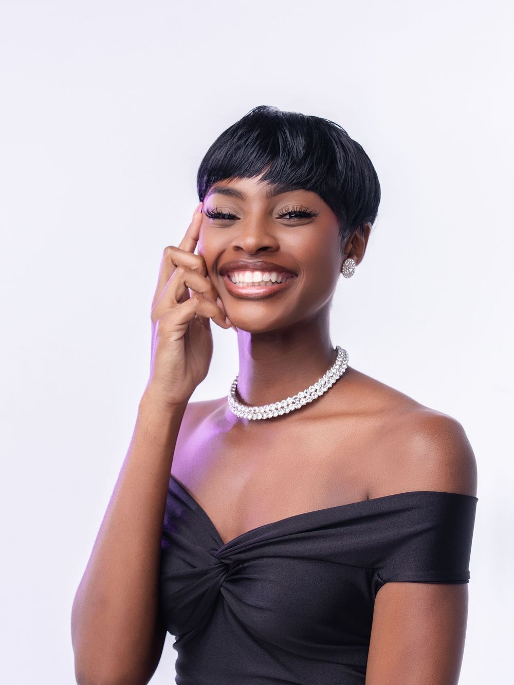 Smiling young woman in black off-shoulder dress with pearl necklace and short hair posing against white background.