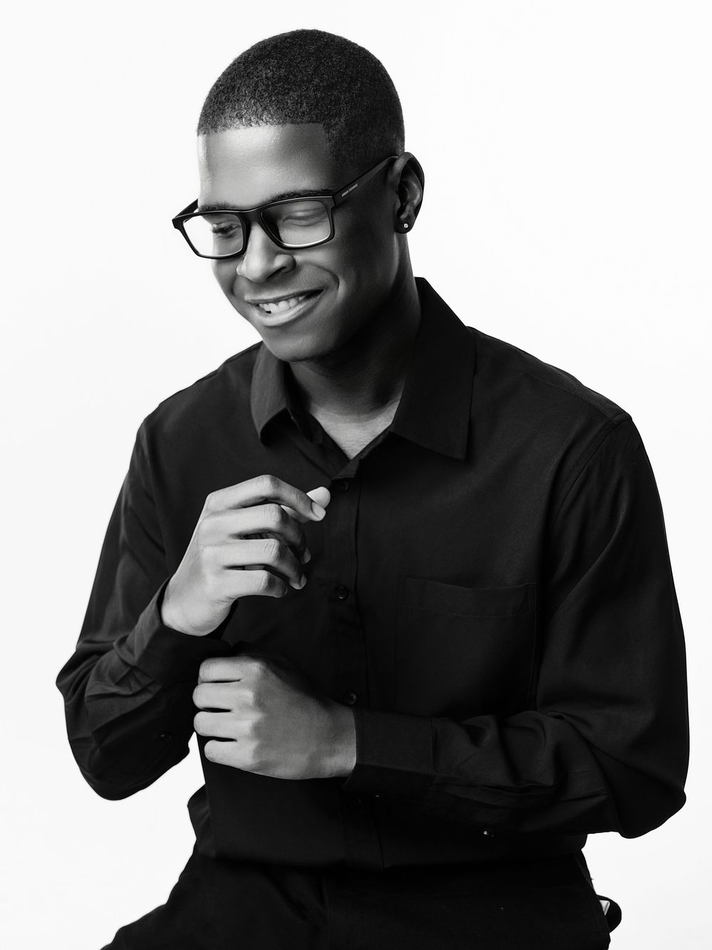 Smiling young man in black shirt and glasses poses confidently in a black and white portrait photo.