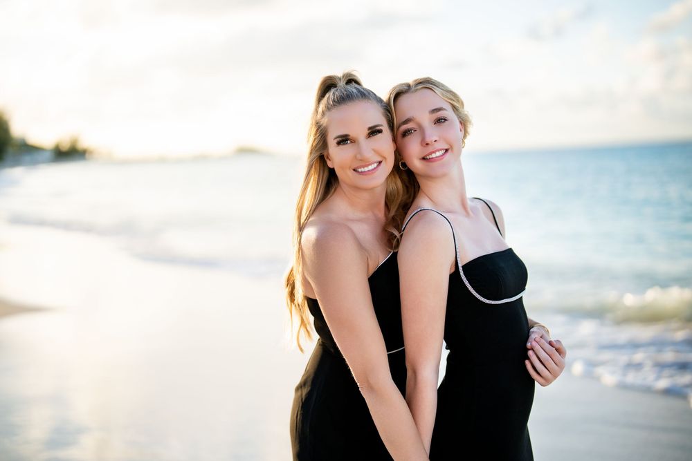 Two people in black dresses pose together on tropical beach at sunset.