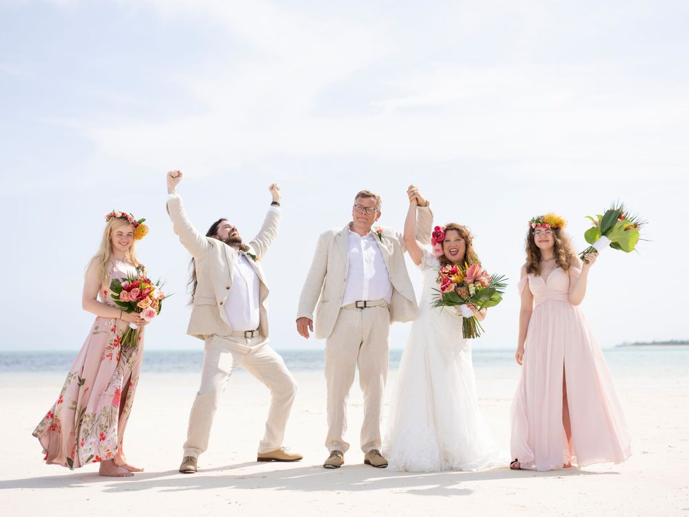 Joyful wedding party celebrating on a sunny beach, bride in white, bridesmaids in floral and pink dresses with flower crowns.