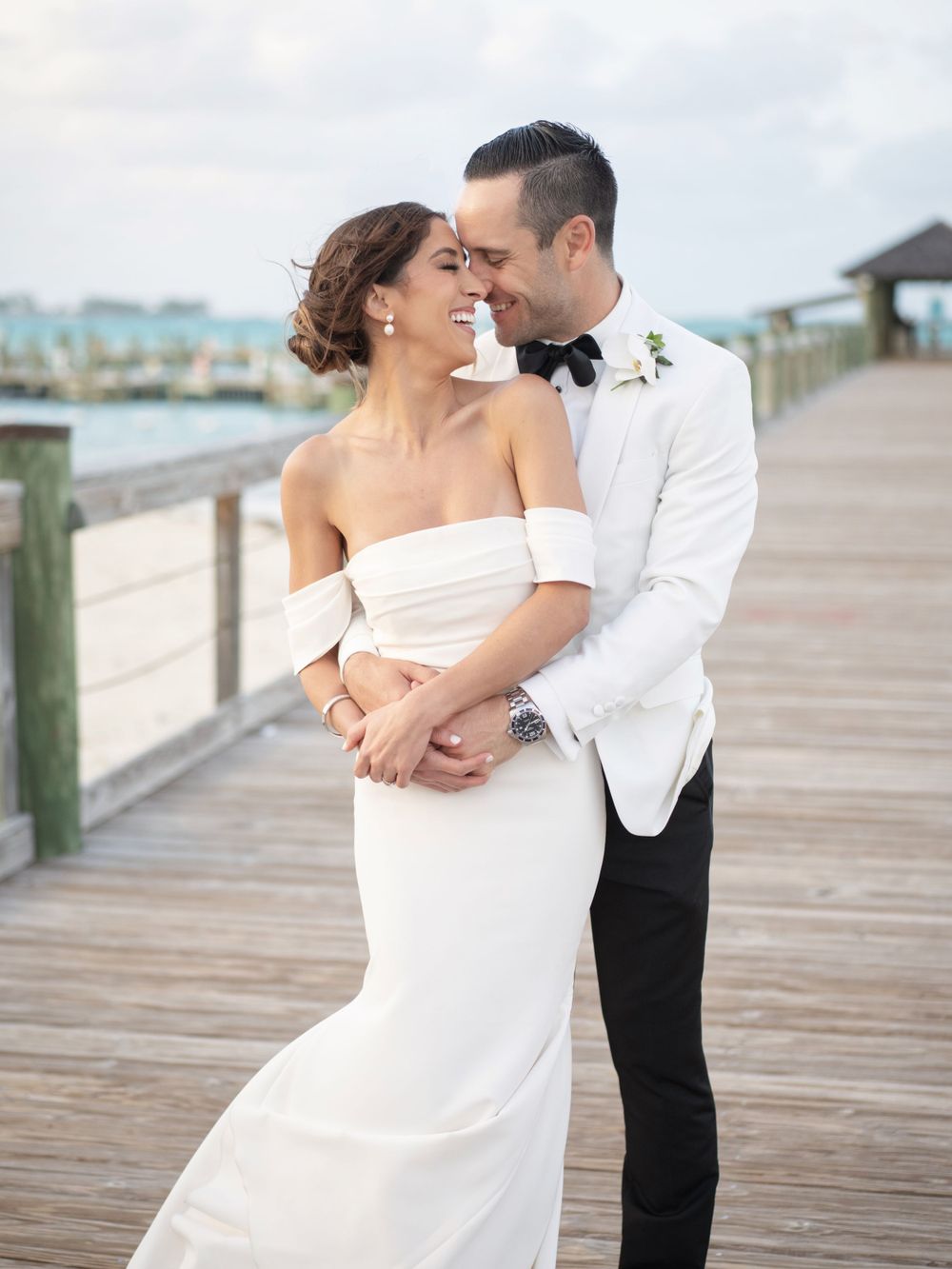Newlywed couple embraces on wooden boardwalk by the water.