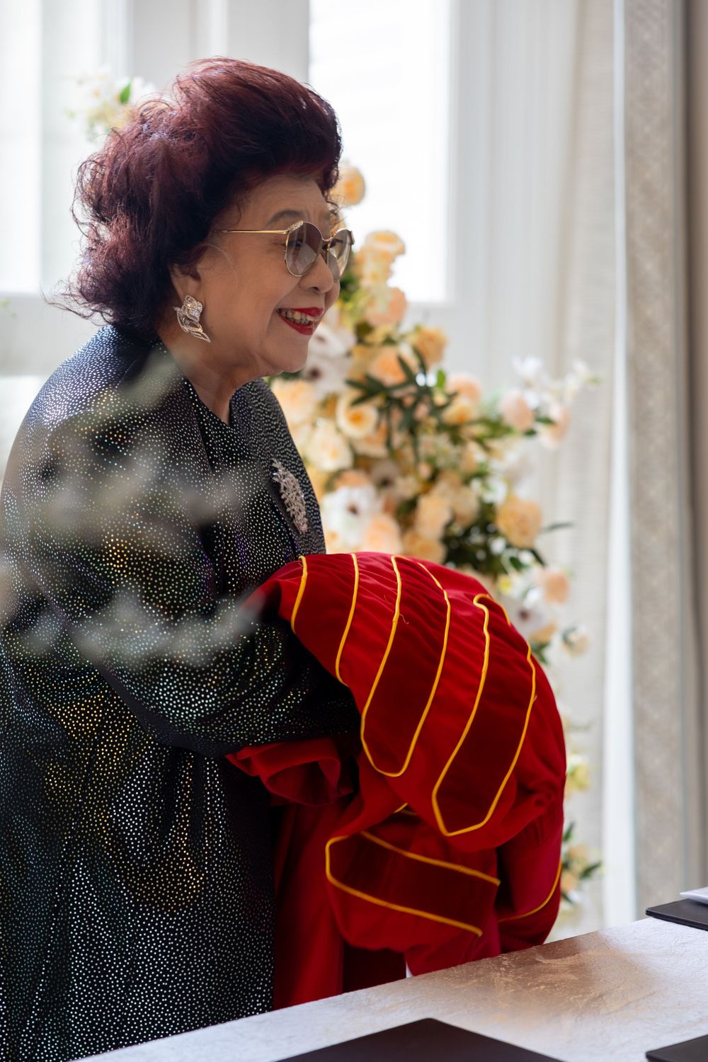 A woman in a dark dress holds a red ceremonial object while standing near floral arrangements.