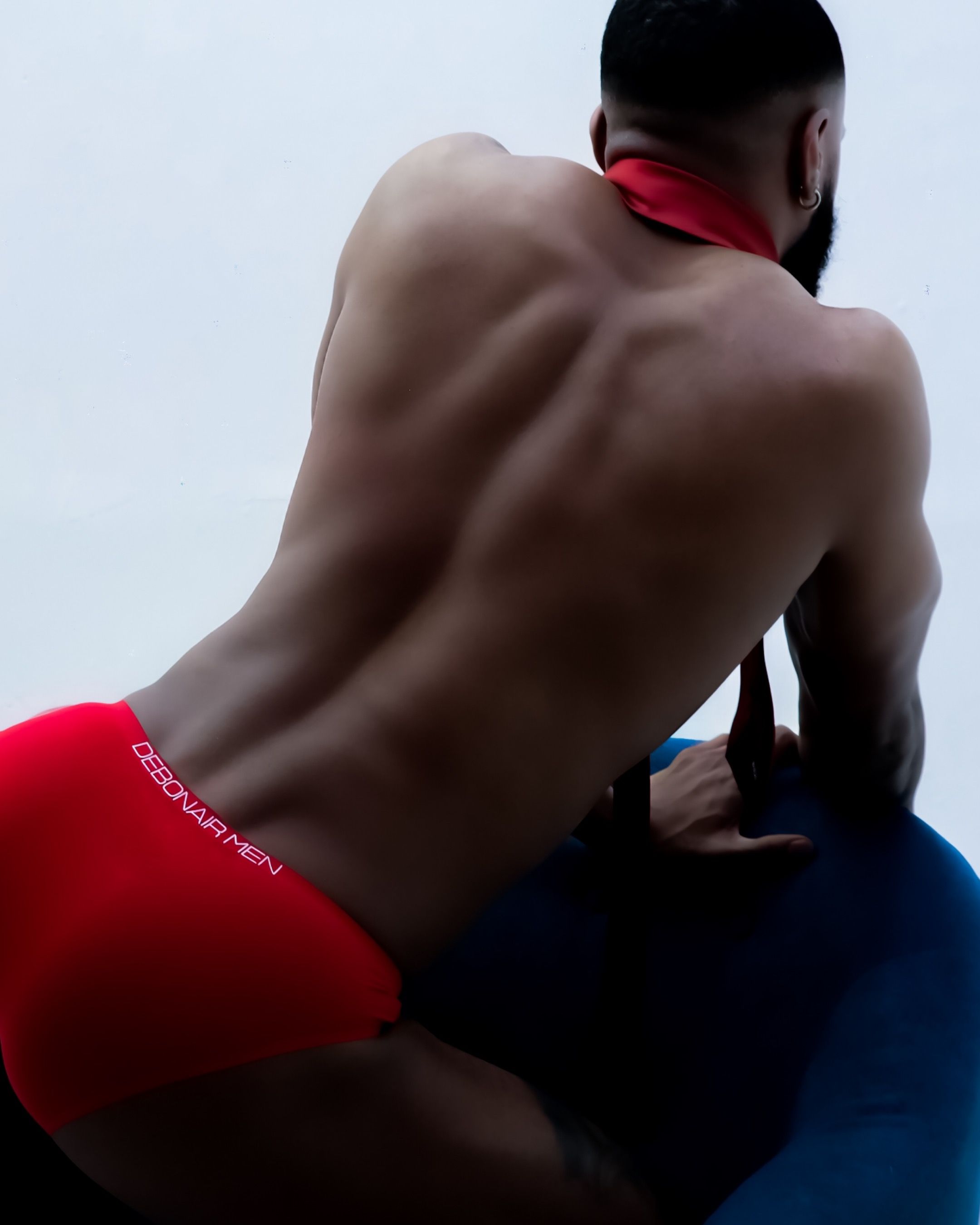Athletic person wearing red briefs sits on blue surface showing muscular back in artistic portrait photography.