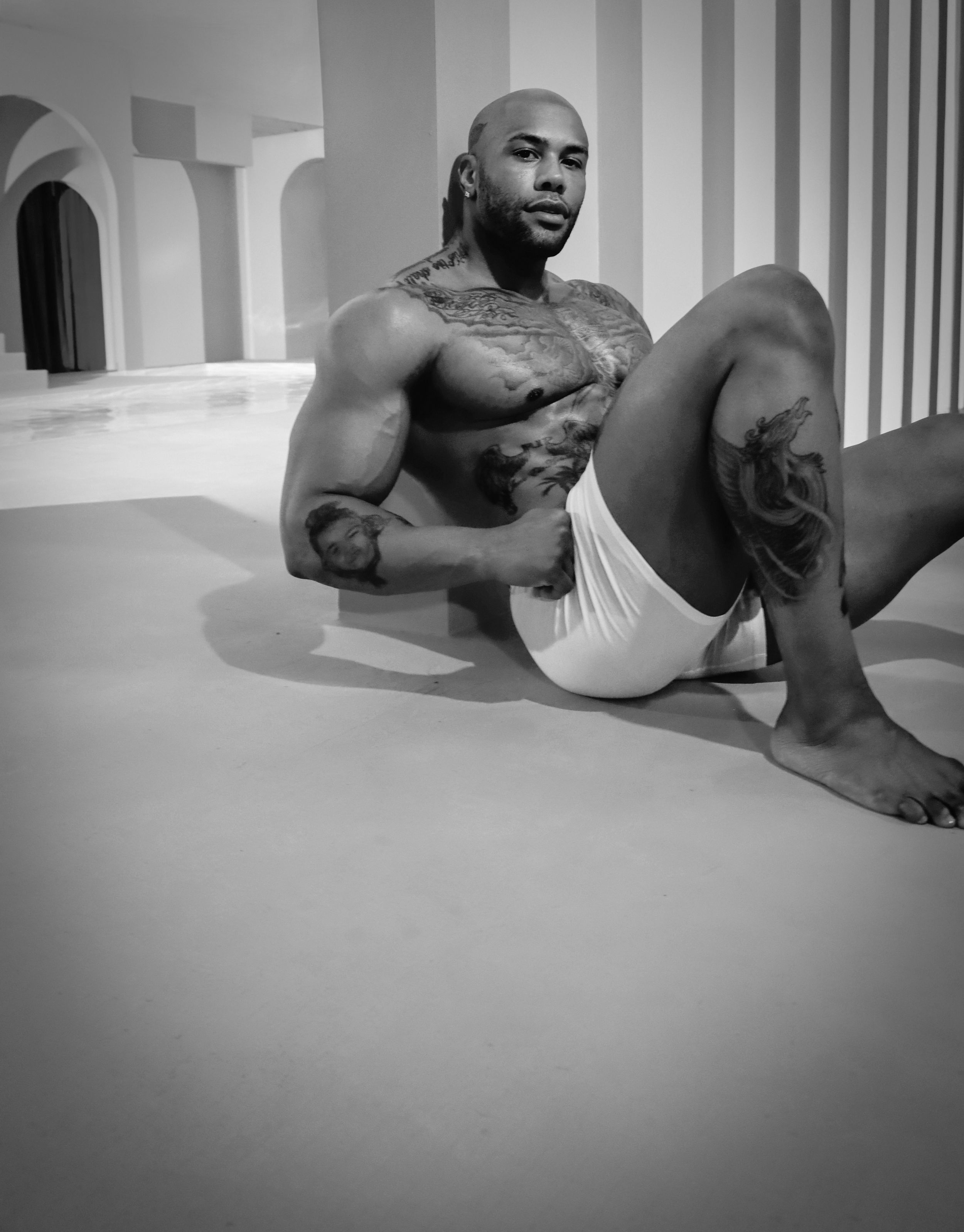 A muscular and tattooed man in white shorts reclining against a white architectural backdrop in black and white.