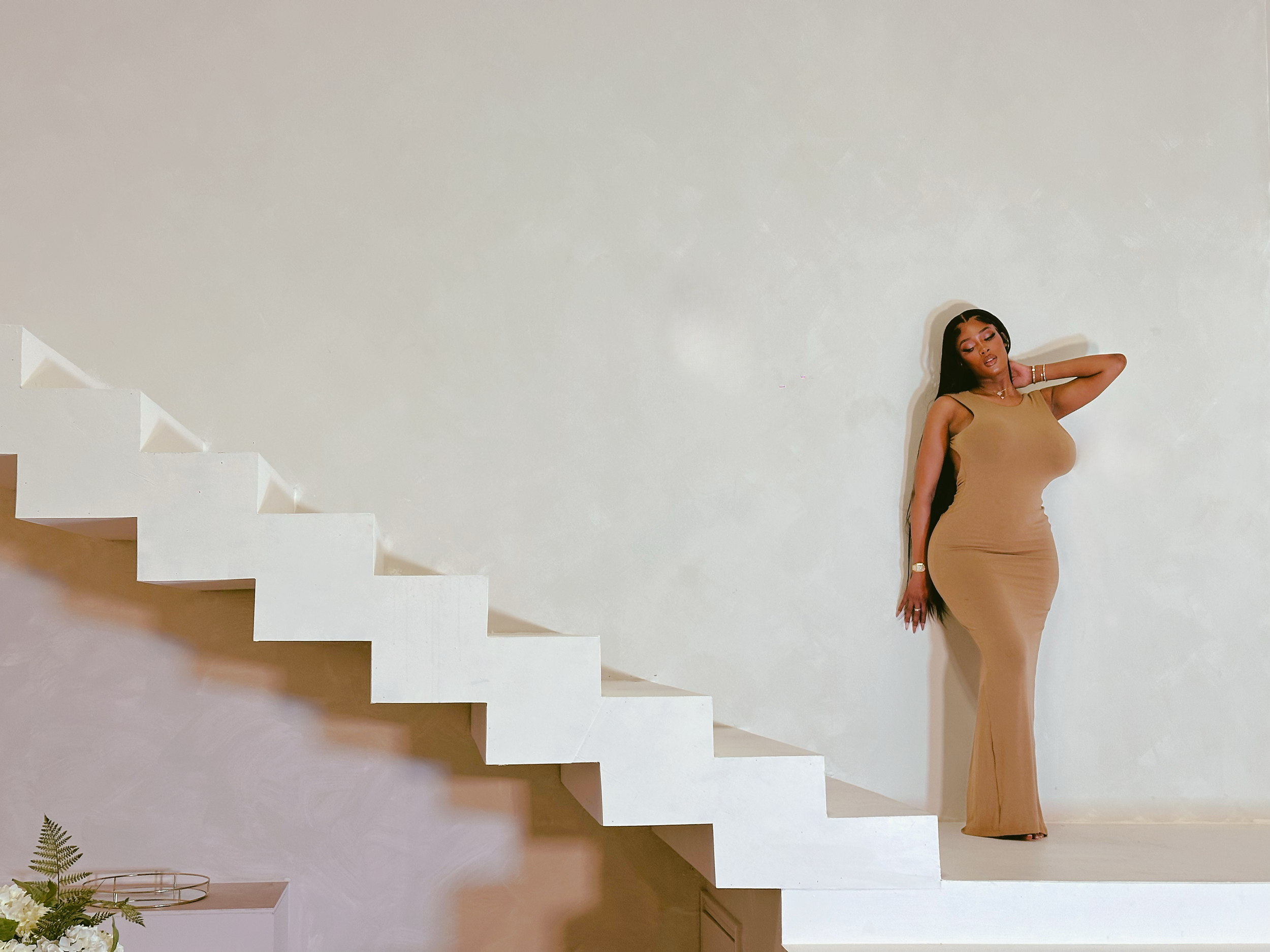 Figure in a beige dress poses elegantly against a white wall with modern angular staircase in contemporary minimalist setting.
