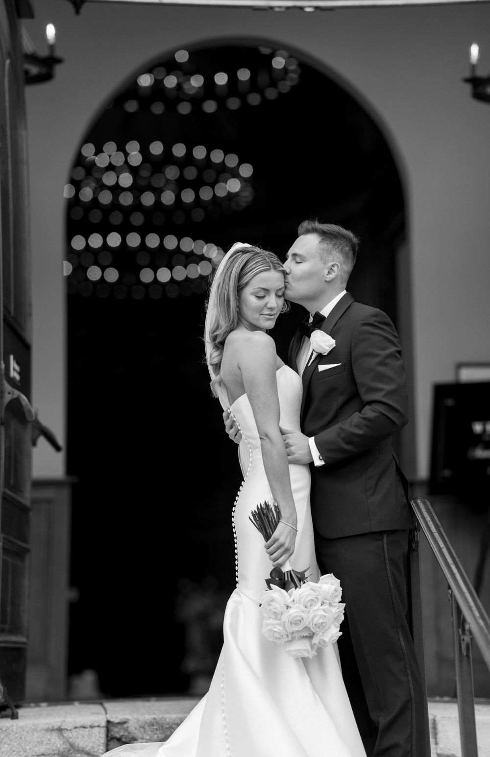 Black and white wedding photograph of a couple embracing under an archway with decorative string lights.