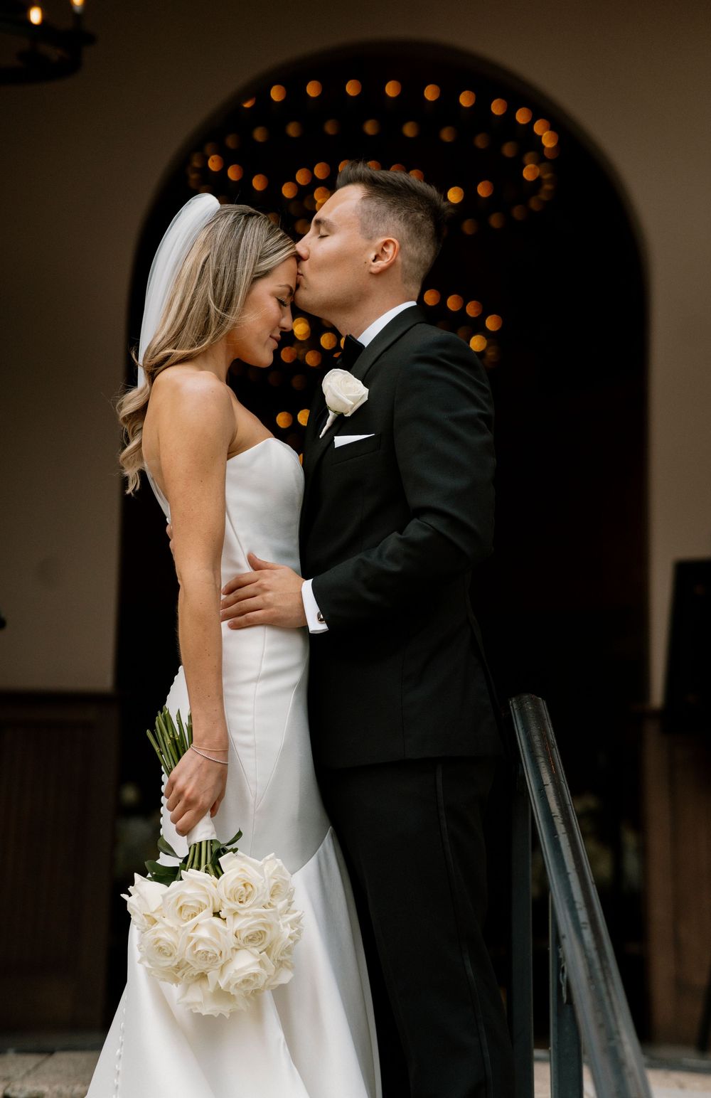 An intimate wedding moment captured beneath an archway decorated with glowing lights as couple shares a tender kiss.
