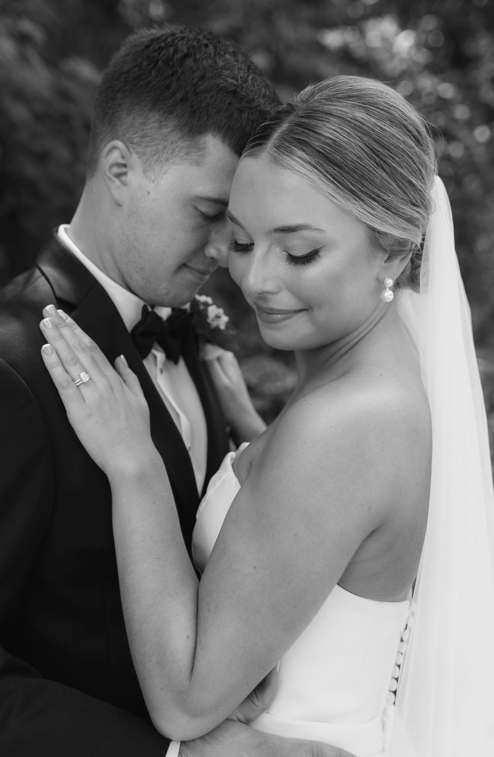 Black and white intimate wedding portrait of newlyweds sharing a tender moment outdoors in formal attire.