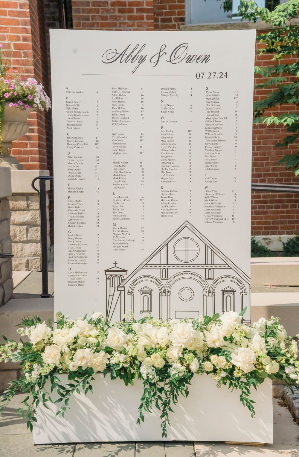 Wedding seating chart with church illustration displayed on white board decorated with lush white floral arrangement.