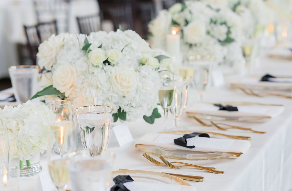 Elegant white wedding reception table setting with floral centerpieces, gold cutlery and glowing candles.