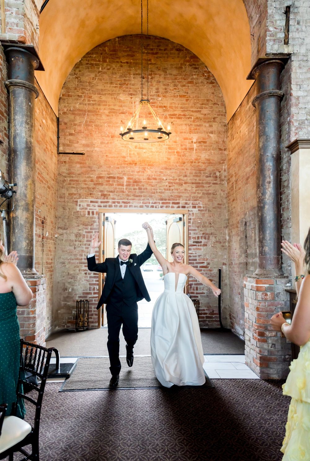 A wedding couple celebrates while walking through a rustic brick archway with decorative columns and an elegant chandelier.