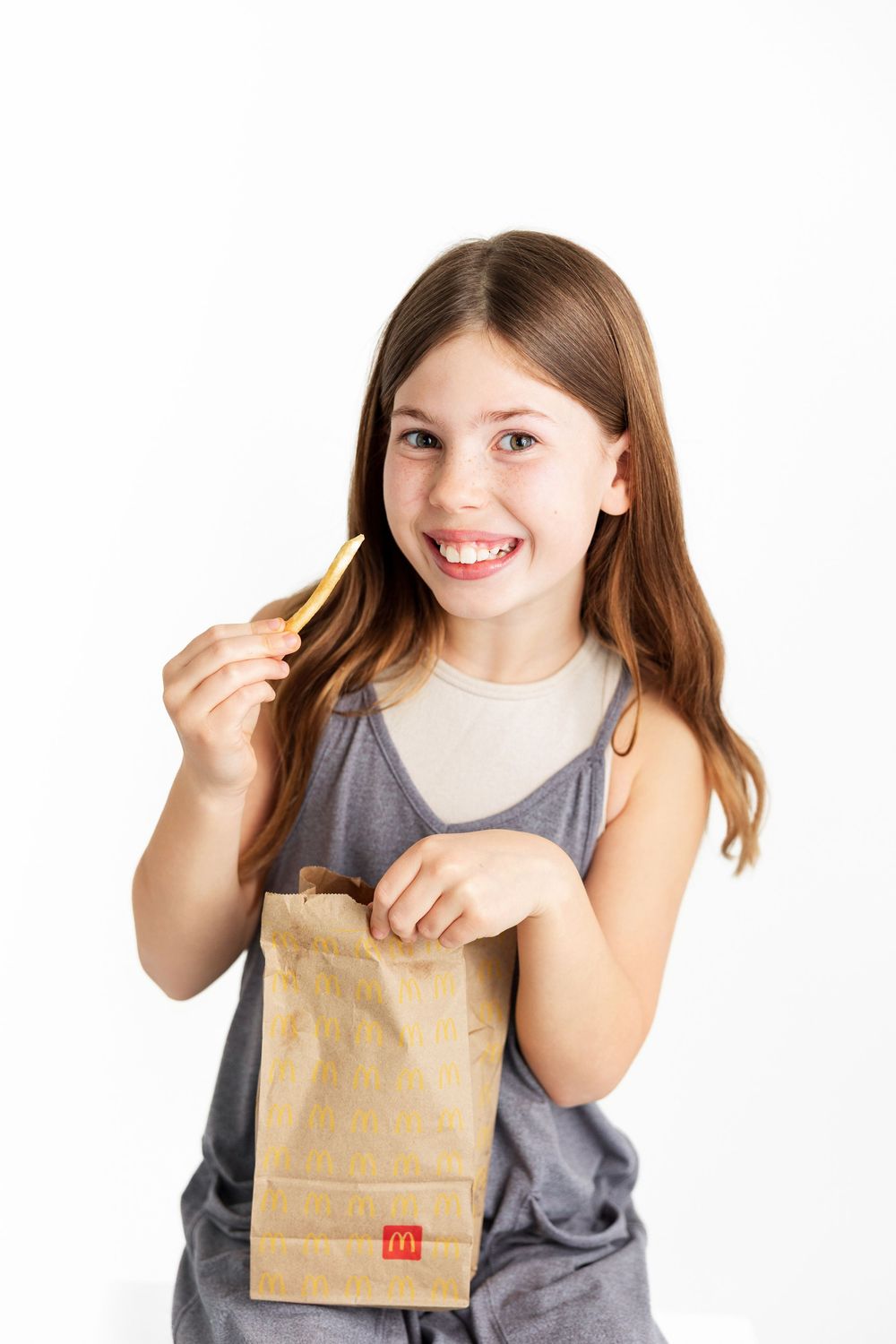A young person in a gray tank top holds a McDonald's paper bag and french fry while smiling against a white background.