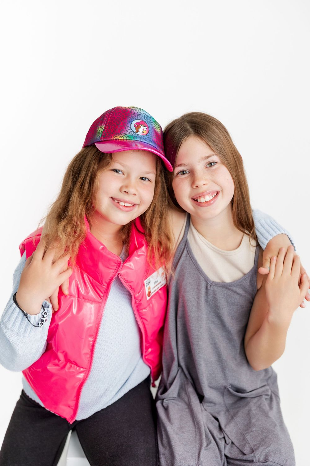 Two young children in casual clothing pose together, one wearing a bright pink vest and baseball cap against white background.