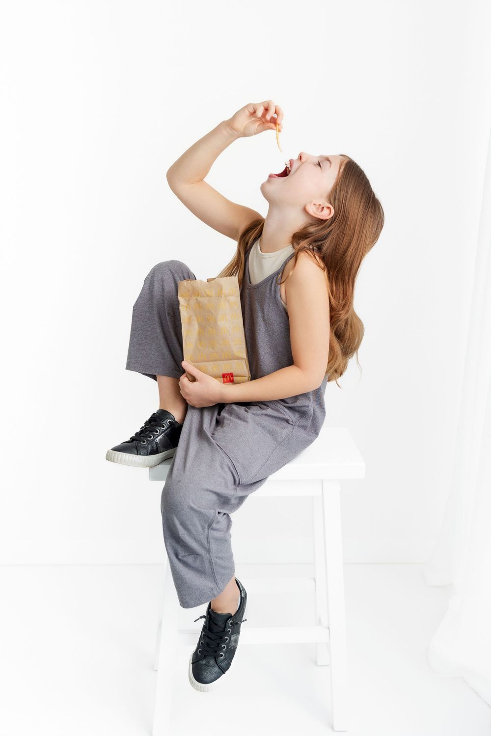 Child in gray jumpsuit sitting on white stool laughing with brown paper bag in hand.