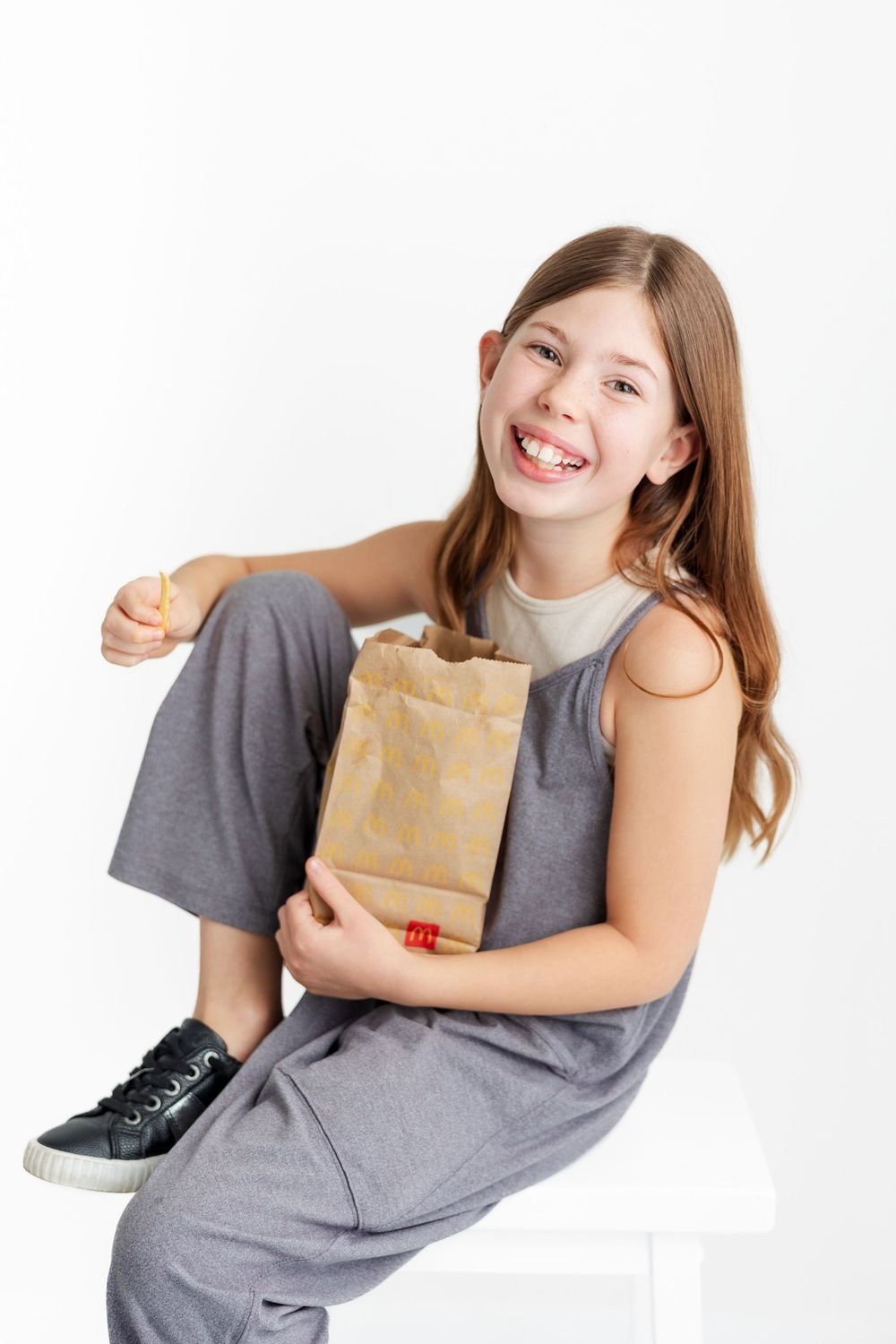 Young person in grey jumpsuit smiling while sitting and holding brown paper bag against white background.