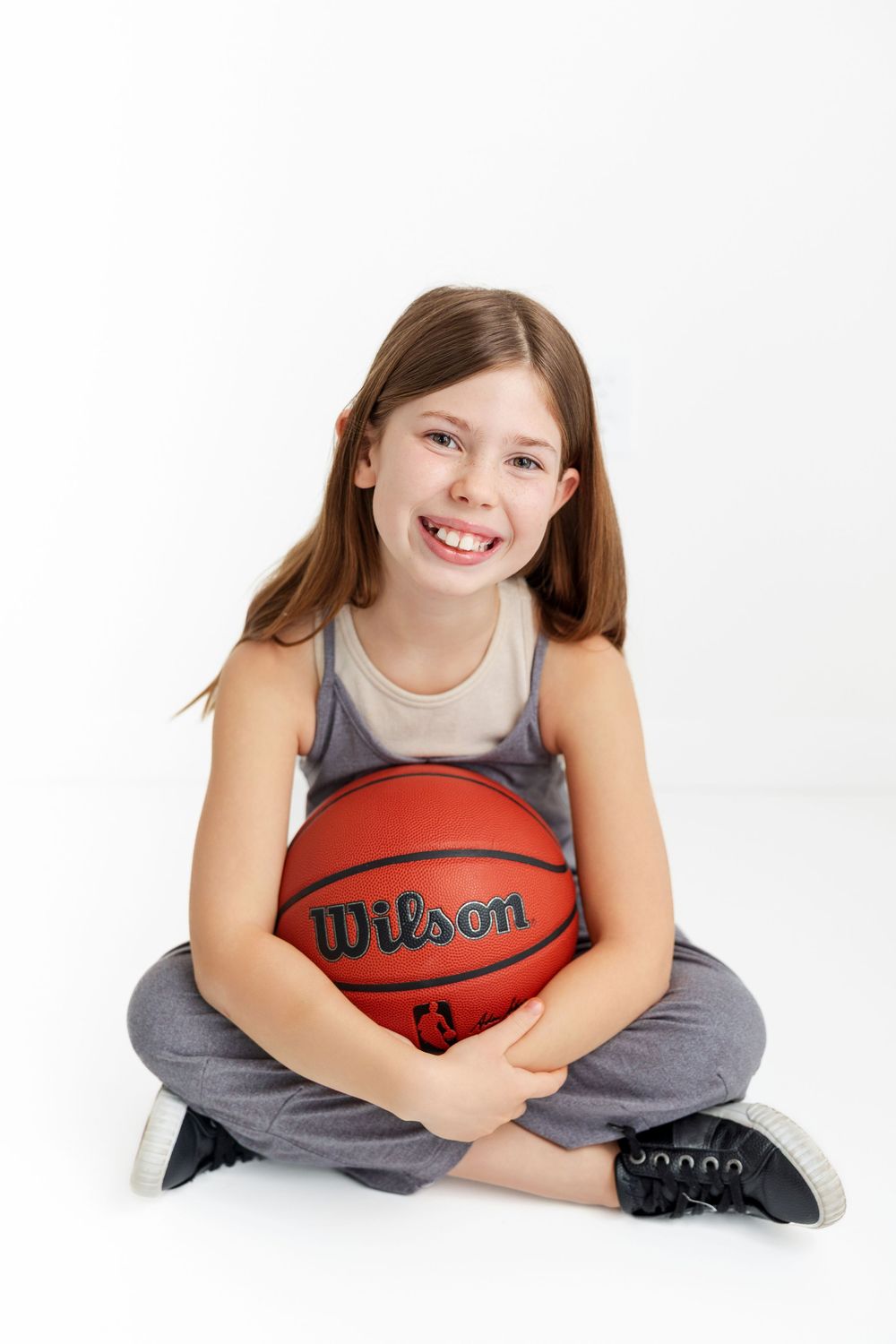 A young person sitting cross-legged in athletic wear holds a Wilson basketball while smiling against a white backdrop.