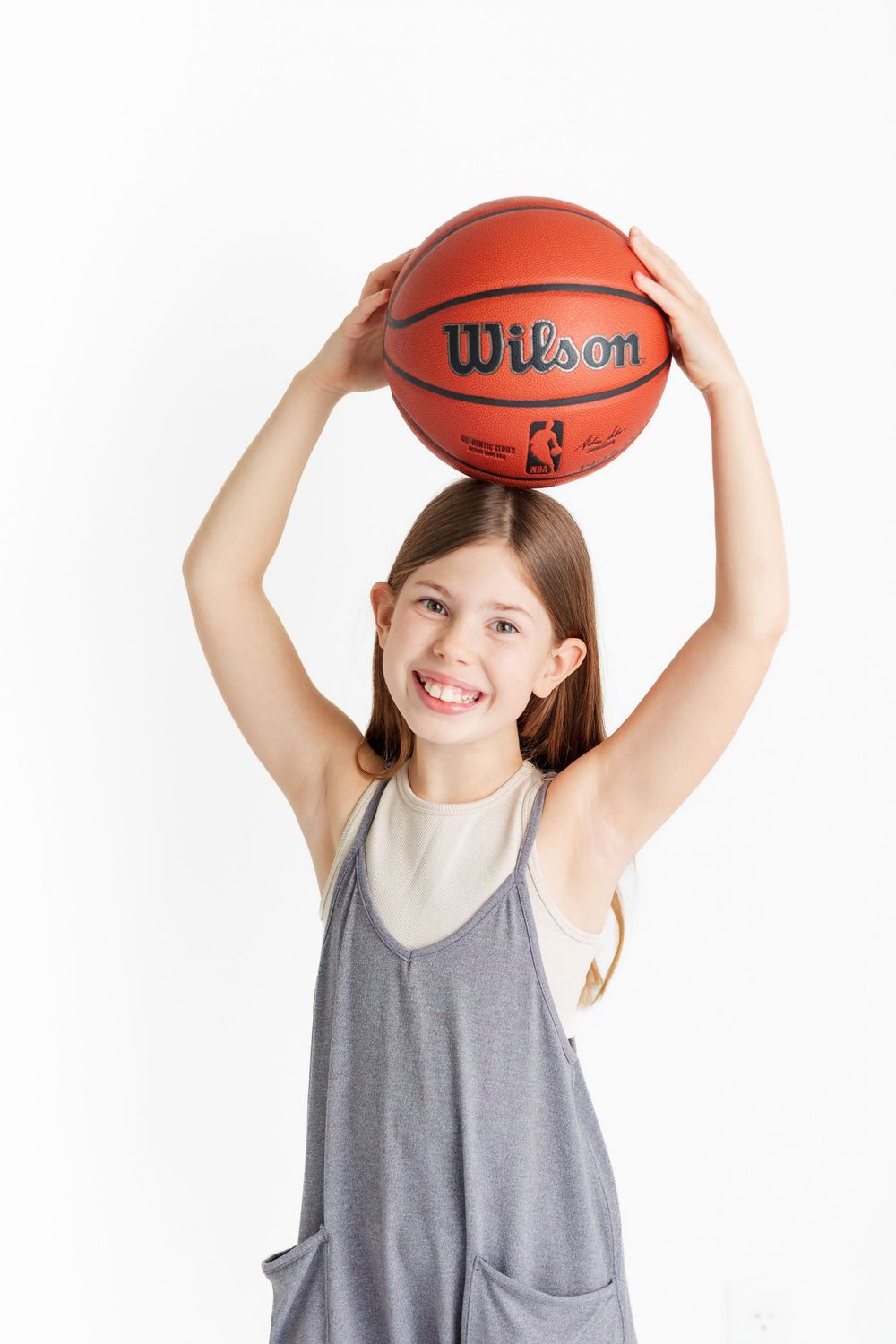 Young kid holding up a Wilson NBA basketball while wearing a gray tank top against white background.