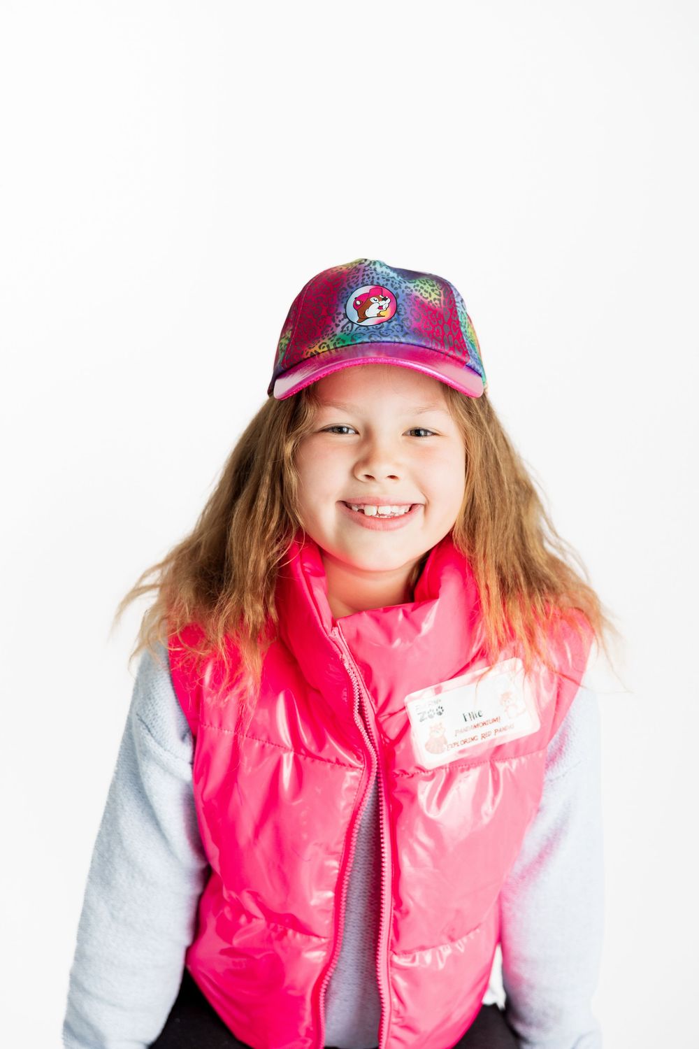 A young person wearing a colorful cap and bright pink puffy vest against a white background.