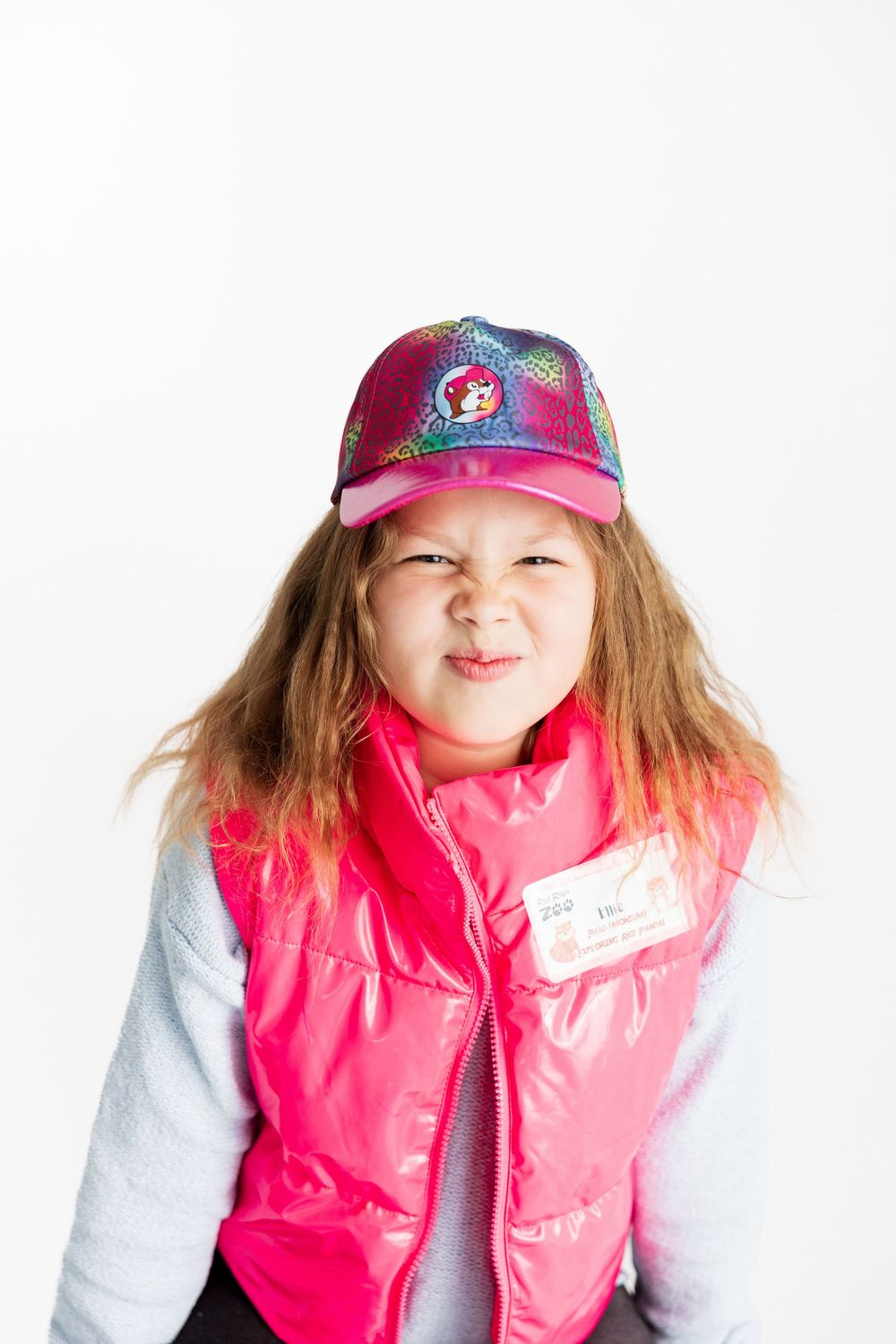 Child wearing pink puffy vest and glittery Disney Princess baseball cap makes playful expression against white background.