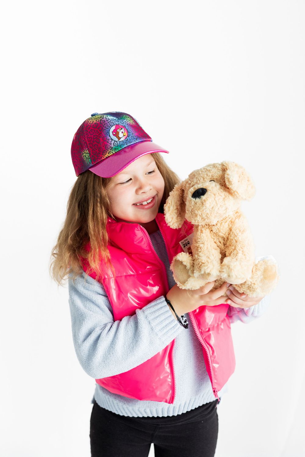 Child wearing a pink vest and baseball cap holds a plush teddy bear against white background.