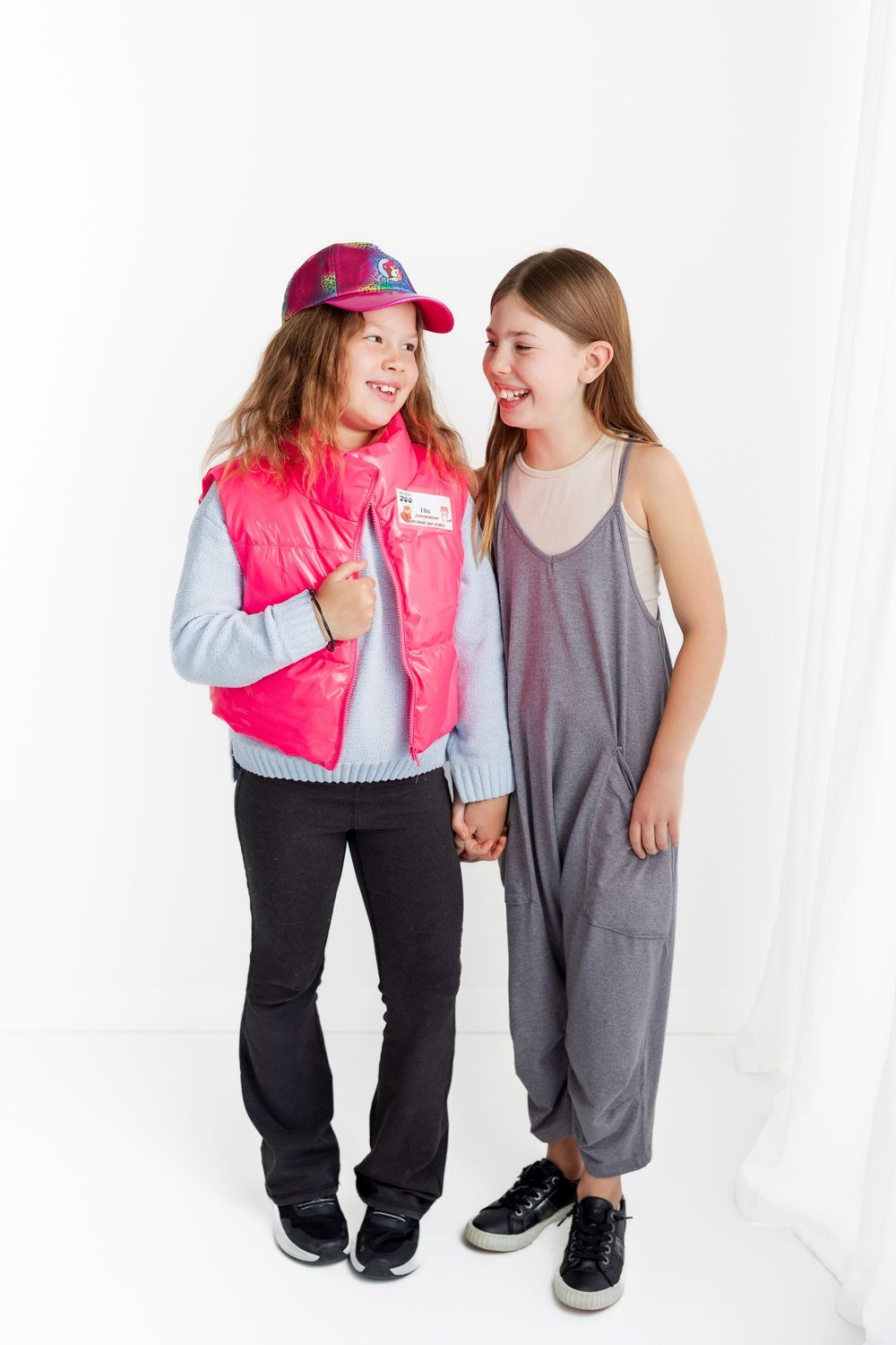 Two children smile and laugh together while one wears a pink vest and baseball cap and the other a gray jumpsuit.