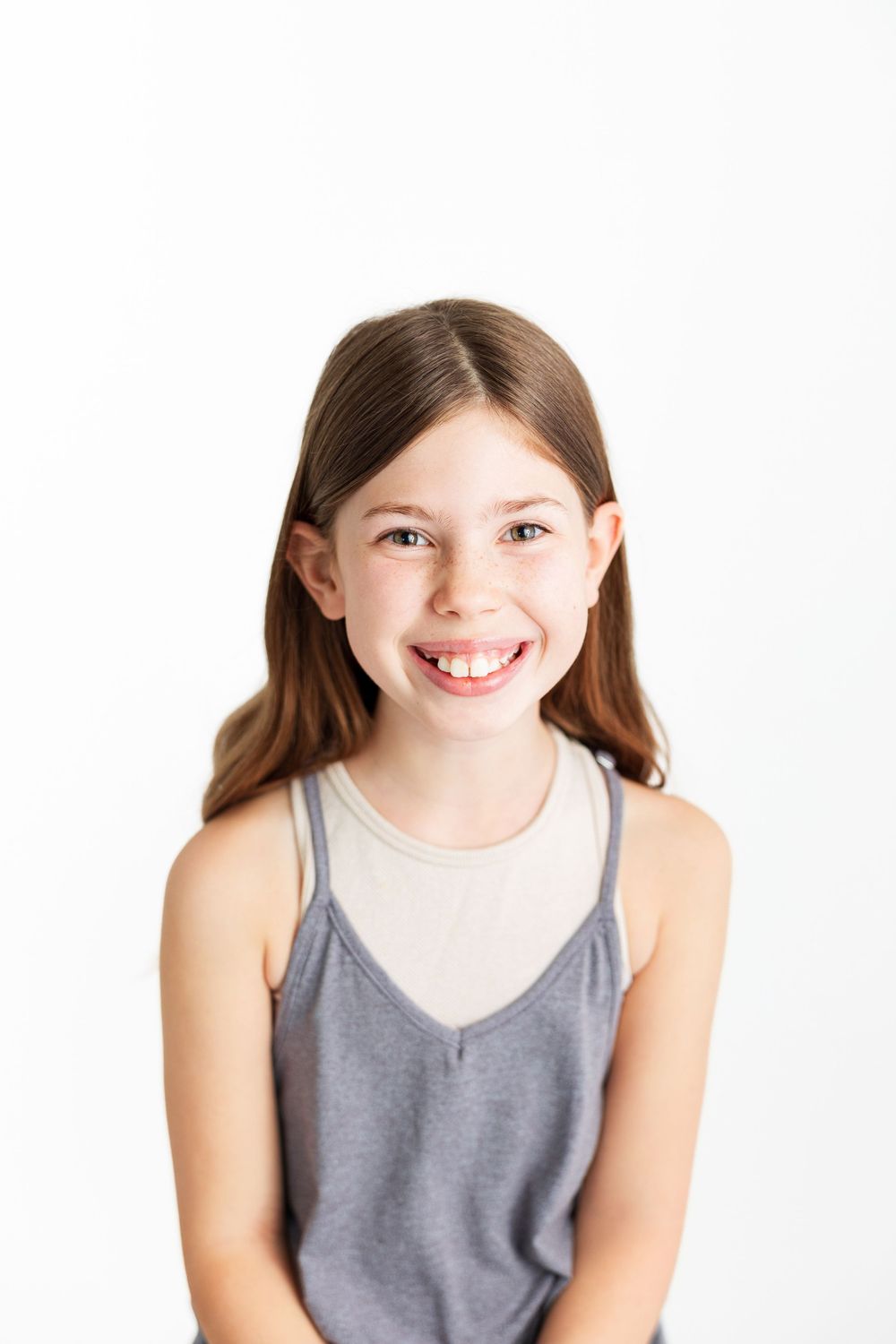 A young person with long brown hair smiles while wearing a gray tank top against a white background.
