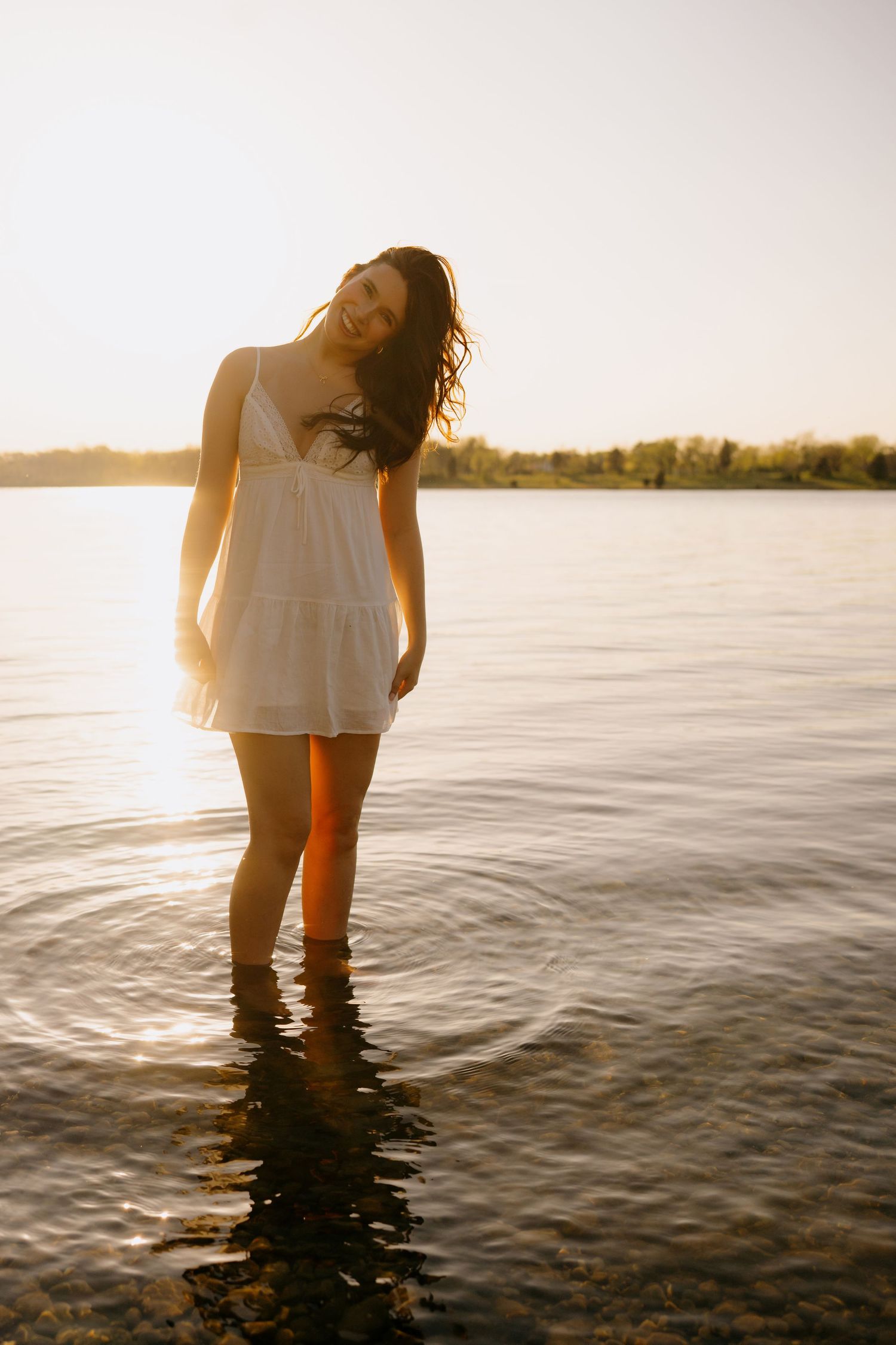 Person in white summer dress standing in shallow water during golden sunset at peaceful lake.