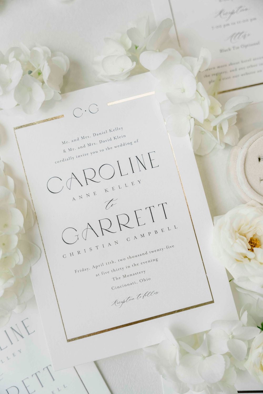 An elegant white wedding invitation with gold text for Caroline and Garrett surrounded by delicate white flower petals.