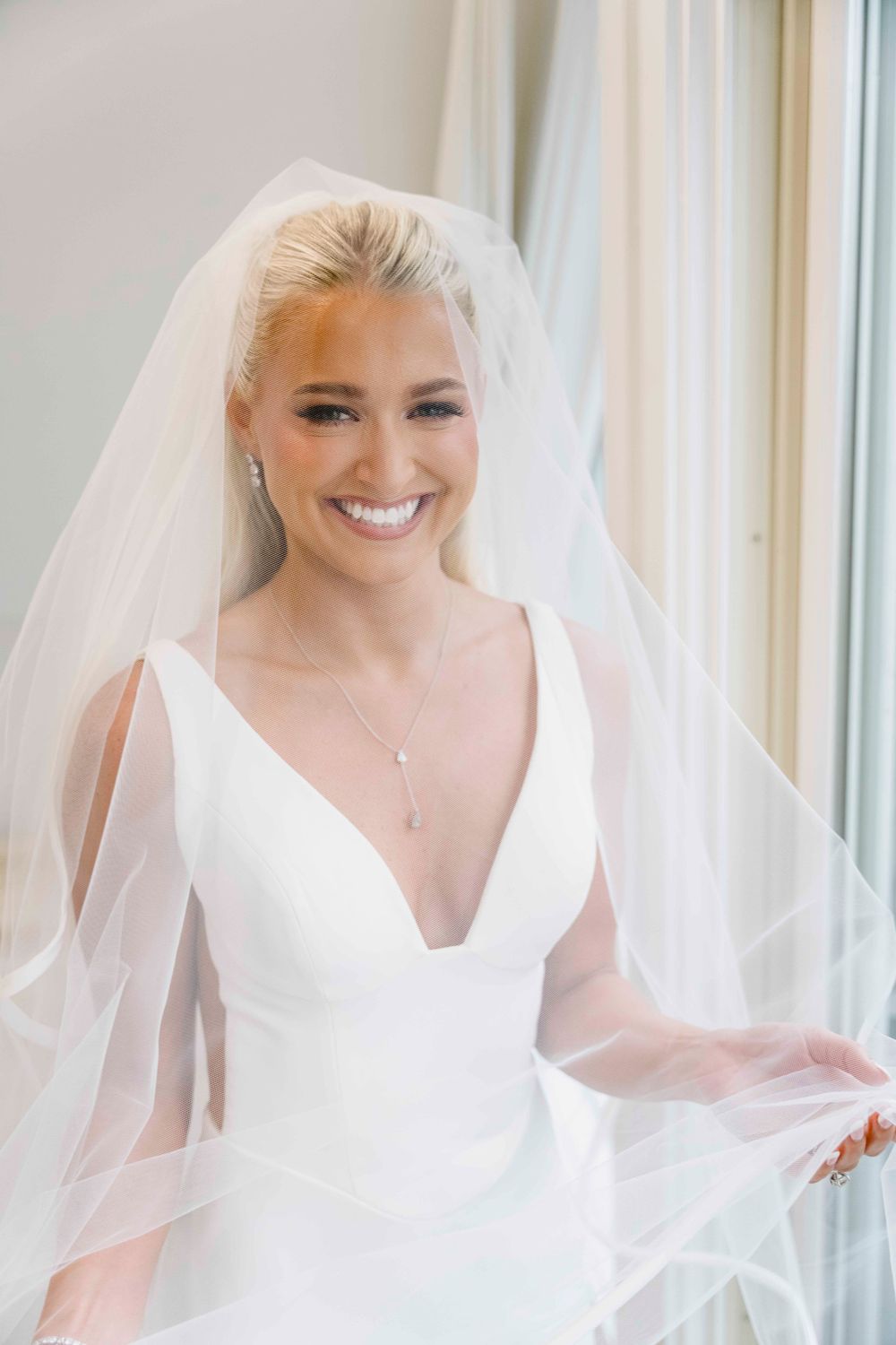 Elegant bride wearing a simple white v-neck wedding gown and long veil poses by a window with natural light.