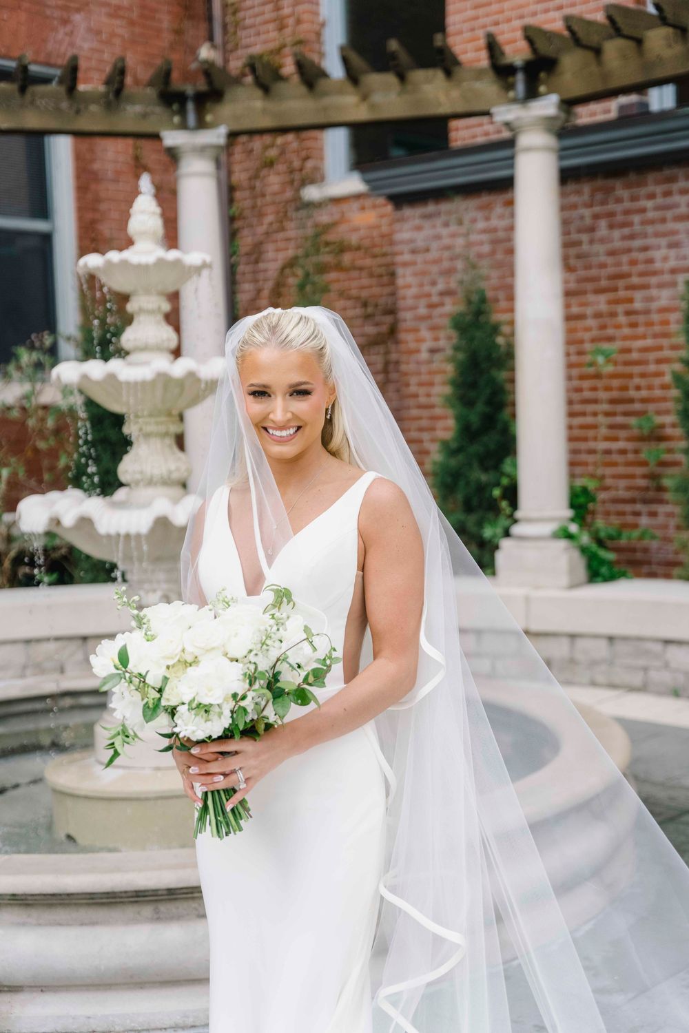 A bride in an elegant white sheath dress and flowing veil holds a white floral bouquet near a brick mansion and fountain.