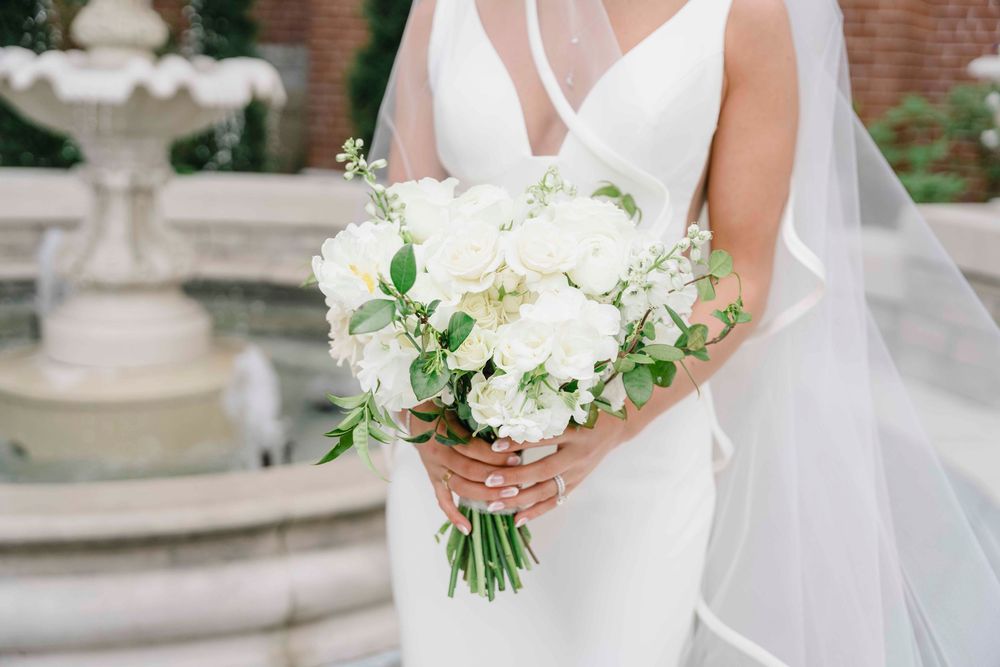 Beautiful white wedding bouquet with roses and greenery held against elegant white bridal gown near stone fountain.