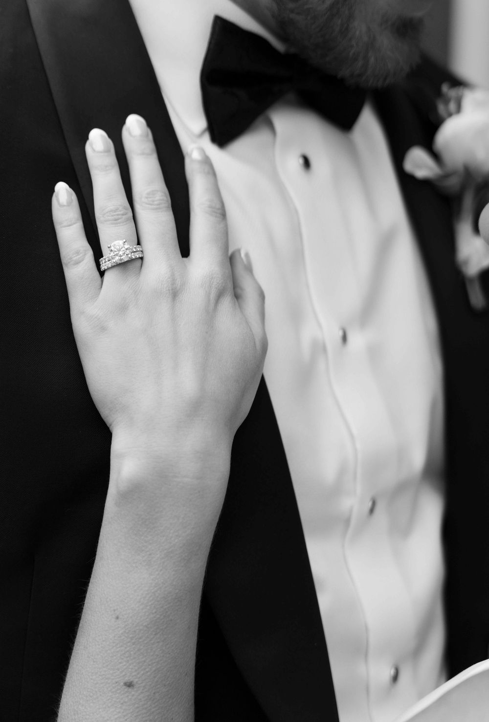 A black and white close-up of a diamond engagement ring on a hand against formal wedding attire.