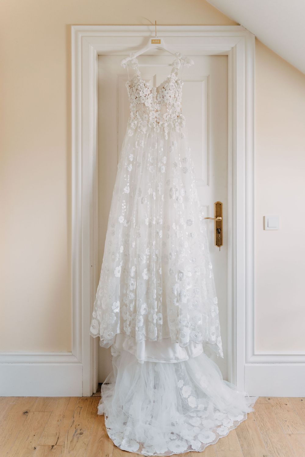 Elegant white lace wedding dress with floral details hanging on white wooden door in natural light.