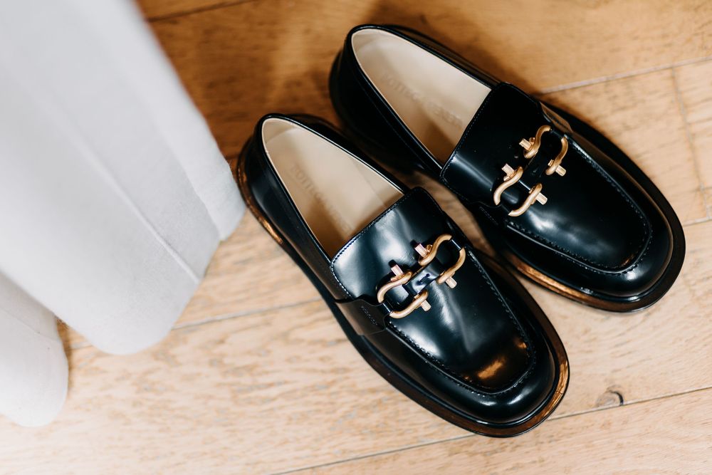 A pair of black patent leather loafers with gold buckle details sits on a wooden surface beside white fabric.