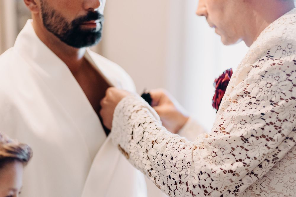 A person helps adjust a white suit jacket and tie during a formal dressing preparation.
