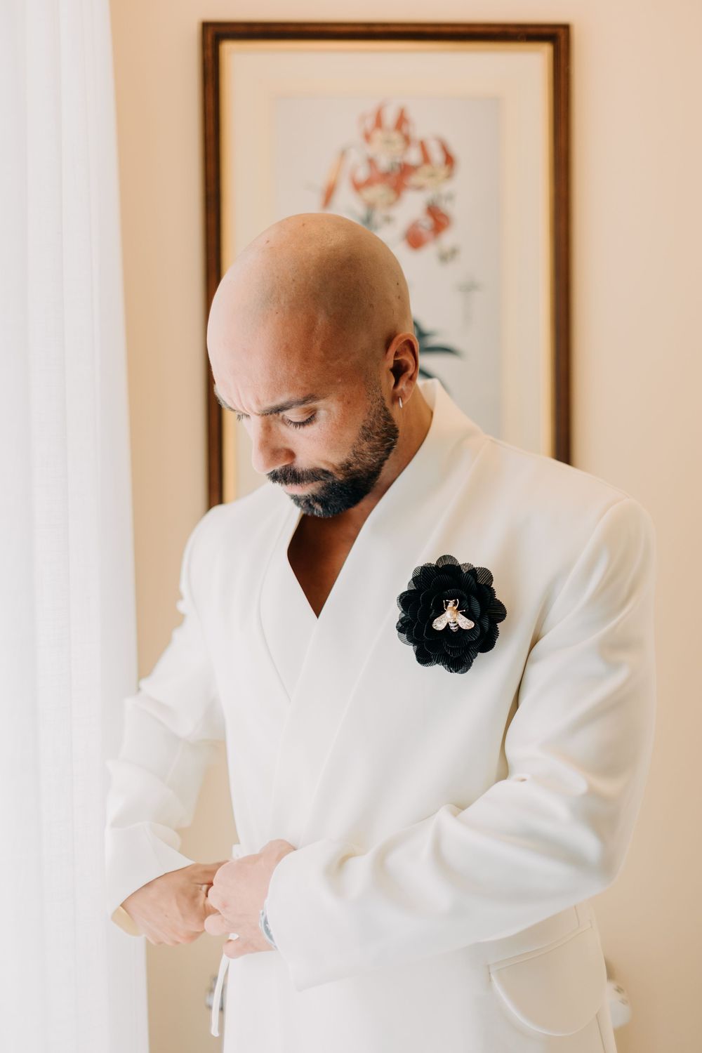 Elegant groom in crisp white tuxedo with black floral boutonniere adjusts his cuff while looking down thoughtfully.