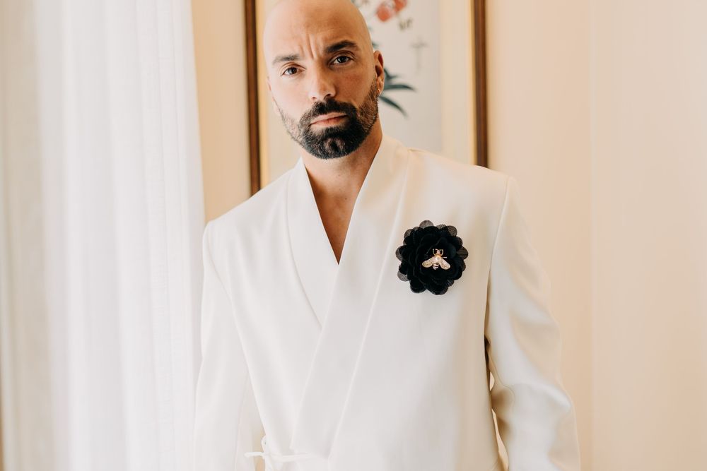 A person wearing a crisp white suit jacket with a black floral boutonniere poses against a light colored wall.