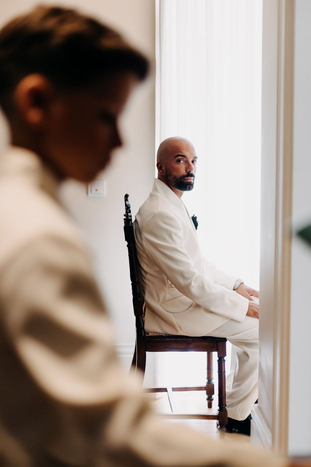 A stylish groom in an all-white tuxedo sits on a dark wooden chair near a bright window in a contemplative pose.