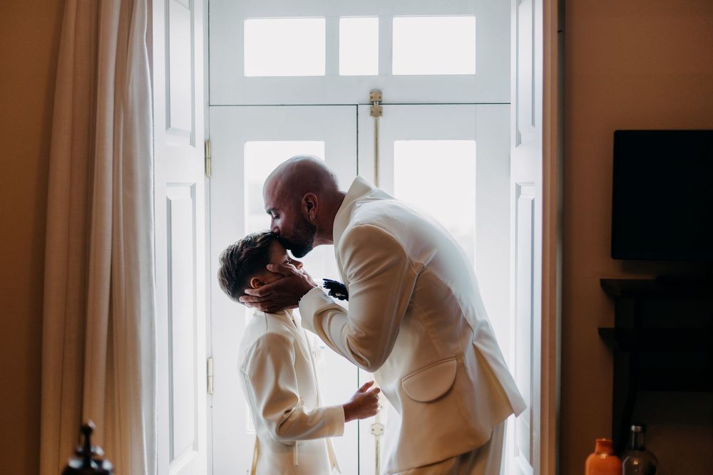 A couple in elegant white attire shares an intimate moment by a sunlit window in their wedding day silhouette.